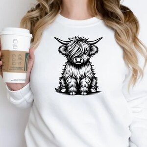 K&ouml;nnte beinhalten: Wei&szlig;es Sweatshirt mit einer Schwarzwei&szlig;-Illustration einer Highland-Kuh mit langem Fell und H&ouml;rnern.
