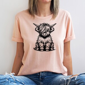 K&ouml;nnte beinhalten: Ein rosa T-Shirt mit einer schwarz-wei&szlig;en Illustration einer sitzenden Highland-Kuh.