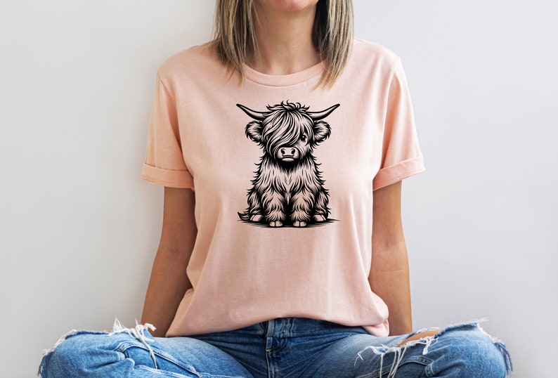 K&ouml;nnte beinhalten: Ein rosa T-Shirt mit einer schwarz-wei&szlig;en Illustration einer sitzenden Highland-Kuh.