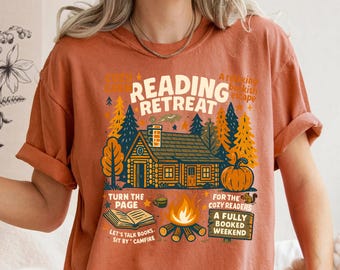 May include: Rust-colored t-shirt with a graphic design featuring a cabin, trees, and a pumpkin. Text includes "Reading Retreat," "Turn the Page," and "A Fully Booked Weekend." The design evokes a cozy, autumnal theme.