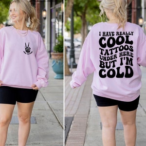May include: Pink sweatshirt with a peace sign and the text "Cool Tattoos" on the front and the text "I have really cool tattoos under here but I'm cold" on the back.