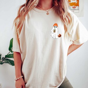 May include: A white t-shirt with a graphic of a ghost wearing an orange beanie and holding a pumpkin and a cup of coffee.