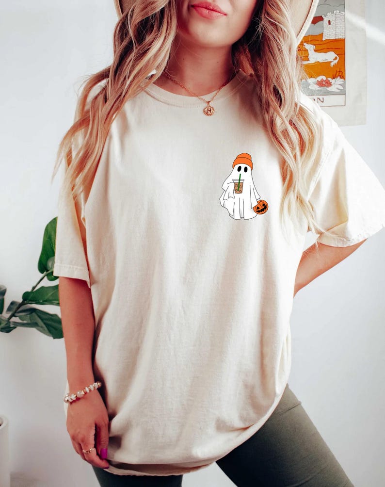 May include: A white t-shirt with a graphic of a ghost wearing an orange beanie and holding a pumpkin and a cup of coffee.