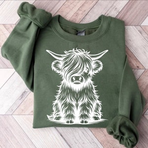 K&ouml;nnte beinhalten: Ein gr&uuml;nes Sweatshirt mit einem wei&szlig;en Aufdruck einer schottischen Hochlandkuh. Die Kuh sitzt und hat ein flauschiges Fell.