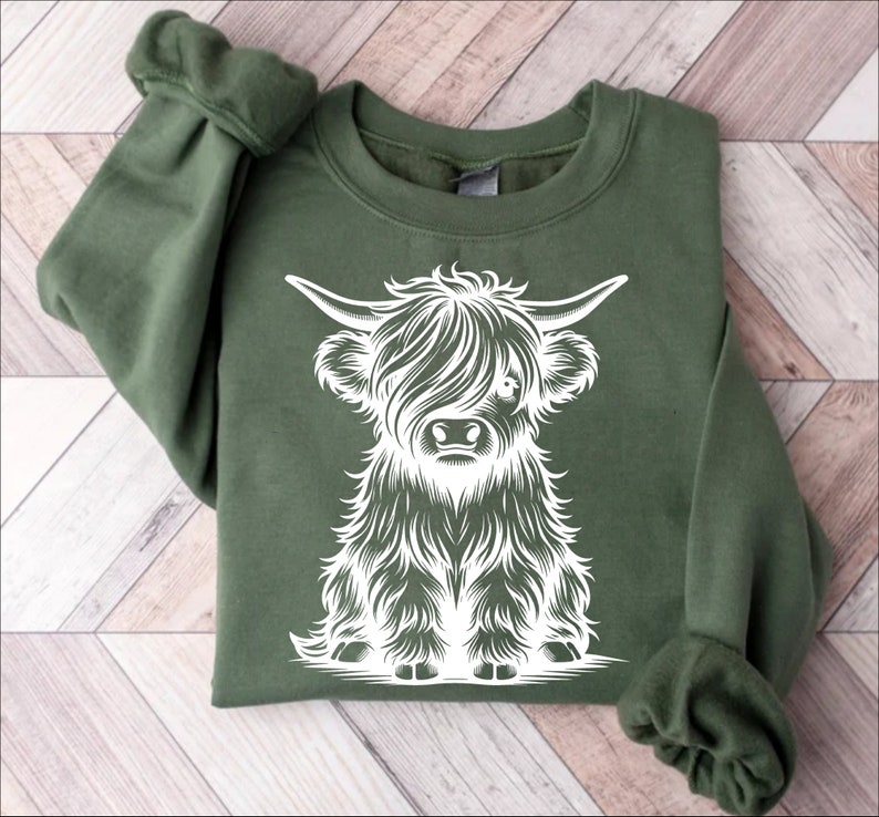 K&ouml;nnte beinhalten: Ein gr&uuml;nes Sweatshirt mit einem wei&szlig;en Aufdruck einer schottischen Hochlandkuh. Die Kuh sitzt und hat ein flauschiges Fell.