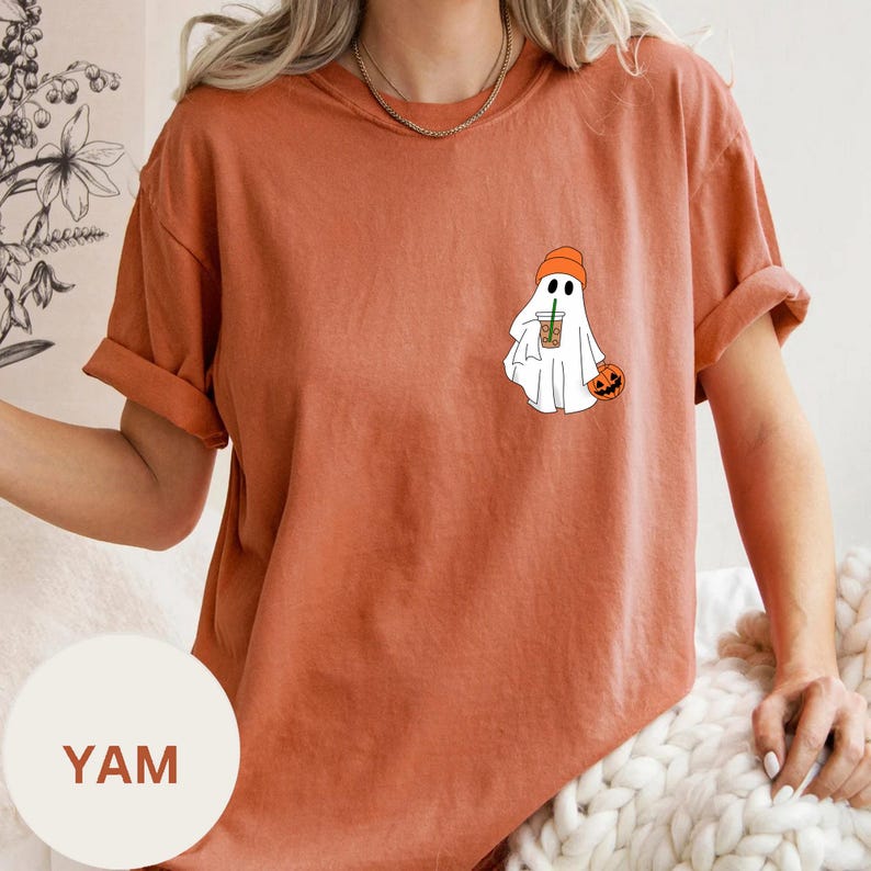 May include: A burnt orange t-shirt with a white ghost wearing a beanie holding a pumpkin and a cup of coffee. The text "YAM" is on a white circle.