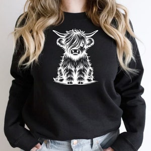 K&ouml;nnte beinhalten: Ein schwarzes Sweatshirt mit einem wei&szlig;en Grafikdruck einer Highland-Kuh mit langem, struppigem Fell. Die Kuh sitzt und schaut den Betrachter an.