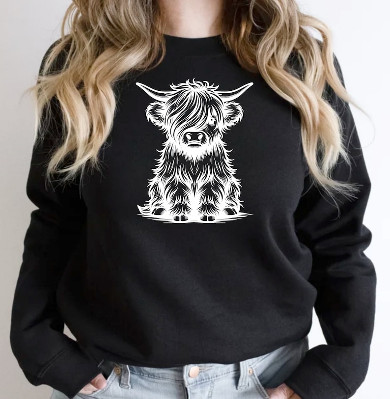 K&ouml;nnte beinhalten: Ein schwarzes Sweatshirt mit einem wei&szlig;en Grafikdruck einer Highland-Kuh mit langem, struppigem Fell. Die Kuh sitzt und schaut den Betrachter an.