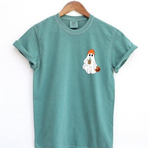 May include: A teal blue t-shirt with a white ghost wearing an orange beanie and holding a pumpkin. The ghost is holding a cup of coffee with a straw.