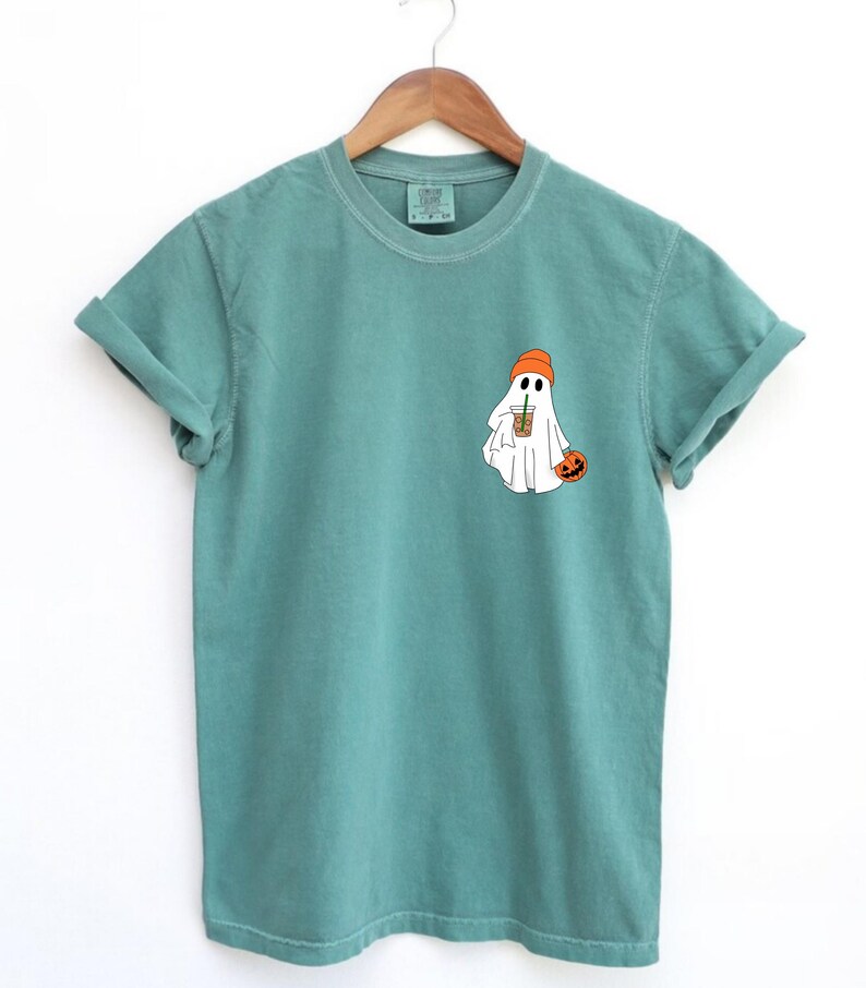 May include: A teal blue t-shirt with a white ghost wearing an orange beanie and holding a pumpkin. The ghost is holding a cup of coffee with a straw.