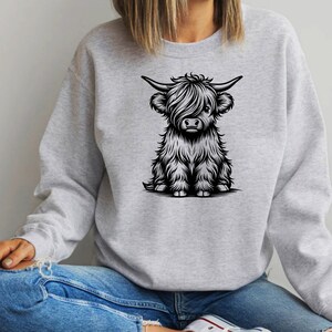 K&ouml;nnte beinhalten: Ein graues Sweatshirt mit einer Schwarzwei&szlig;-Illustration einer Highland-Kuh mit flauschigem Fell. Die Kuh sitzt und schaut den Betrachter an.