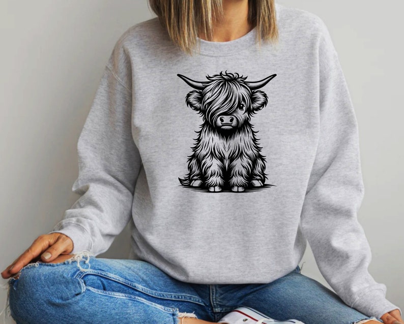K&ouml;nnte beinhalten: Ein graues Sweatshirt mit einer Schwarzwei&szlig;-Illustration einer Highland-Kuh mit flauschigem Fell. Die Kuh sitzt und schaut den Betrachter an.