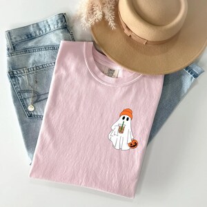 May include: A pink t-shirt with a white ghost wearing an orange hat holding a pumpkin and a cup of coffee. The text "COMFORT COLORS" is visible on the shirt.