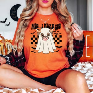 May include: A woman wearing an orange t-shirt with a black and white checkered pattern and a ghost with a cow's head. The shirt says "Moo... I mean Boo".