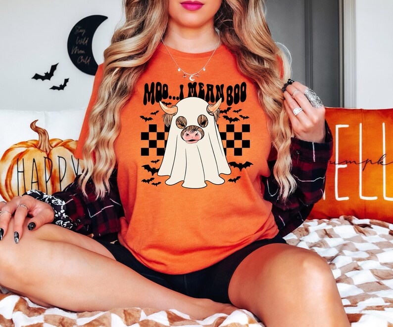 May include: A woman wearing an orange t-shirt with a black and white checkered pattern and a ghost with a cow's head. The shirt says "Moo... I mean Boo".