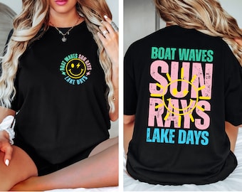 Könnte beinhalten: Schwarzes T-Shirt mit einem grafischen Aufdruck eines Smiley-Gesichts mit Blitzen und dem Text "Boat Waves Sun Rays Lake Days". Die Rückseite des Shirts hat einen größeren grafischen Aufdruck einer Sonne mit dem Text "Boat Waves Sun Rays Lake Days".