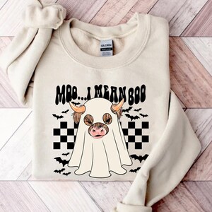 May include: A cream-colored sweatshirt with a black and white checkered pattern and a ghost with a cow's head. The ghost has brown fur and a pink nose. The text "Moo... I mean Boo" is written in black above the ghost.