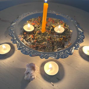 May include: A silver decorative tray with a scalloped edge holds dried herbs and flowers, surrounded by lit tea light candles. A tall yellow candle burns brightly in the center, with a cluster of crystals nearby.
