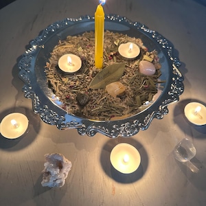 May include: A silver decorative tray with a lit yellow candle in the center, surrounded by small lit tea light candles, crystals, and dried herbs. A bay leaf with the words "Remove Bless" is visible. The arrangement is set on a light-colored surface.