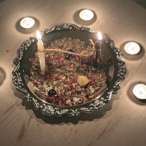 May include: A silver-colored, ornate tray holds two lit candles, dried herbs, and crystals. Surrounding the tray are several lit tea light candles, creating a warm, mystical ambiance. The arrangement is set on a white, distressed wooden surface.