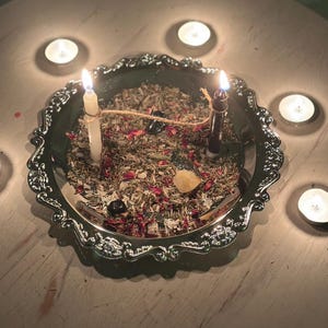 May include: A silver-toned decorative tray filled with dried herbs, flower petals, and small crystals. Two lit candles, one white and one black, are connected by a piece of twine. Four additional tea light candles are arranged in a circle around the tray.