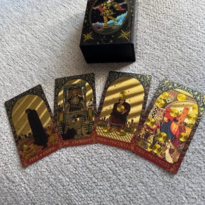 May include: Four tarot cards with gold foil accents and red borders. The cards are: Five of Cups, The Chariot, Four of Pentacles, and Ten of Pentacles. The cards are laid out on a white surface.