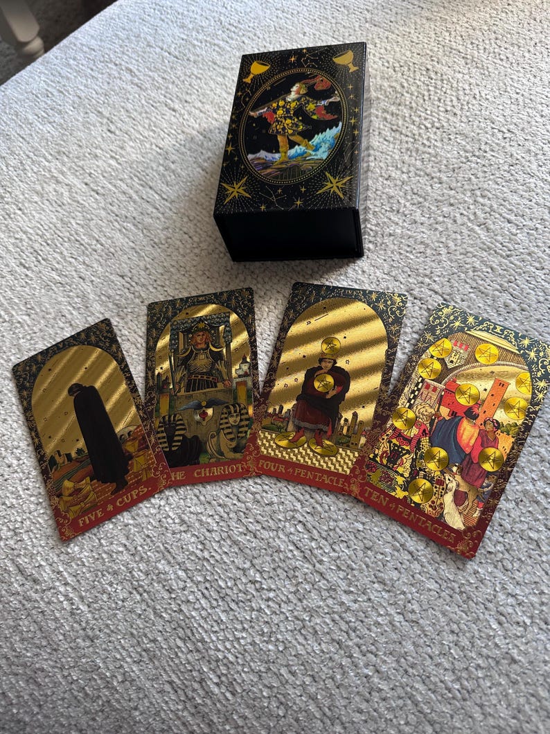 May include: Four tarot cards with gold foil accents and red borders. The cards are: Five of Cups, The Chariot, Four of Pentacles, and Ten of Pentacles. The cards are laid out on a white surface.