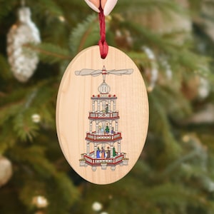Pode incluir: Um enfeite de Natal de madeira em forma oval com uma fita vermelha. O enfeite apresenta uma imagem pintada de uma pirâmide de Natal alemã tradicional com uma hélice giratória no topo.