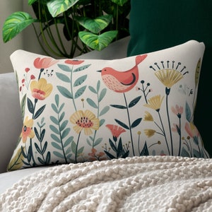 Puede incluir: Una almohada blanca con un diseño floral y de pájaro. La almohada presenta un pájaro rojo con flores amarillas, rosas y verdes sobre un fondo blanco.