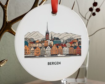 May include: A white round ornament with a colorful illustration of the Bergen, Norway skyline. The ornament has a red ribbon attached to the top and the word "BERGEN" printed in black at the bottom.
