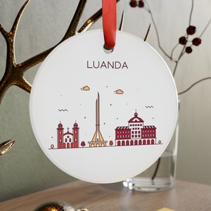 May include: White round ornament with a red ribbon. The ornament features a line drawing of the Luanda skyline in red and yellow. The text "LUANDA" is printed in brown at the top of the ornament.