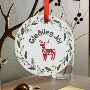 Puede incluir: Adorno navideño redondo blanco con una cinta roja. El adorno presenta una corona verde con bayas rojas y un reno estilizado con un cuerpo estampado en rojo y blanco. El texto "Gleðileg Jól" está escrito en negro dentro de la corona.