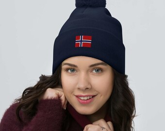 May include: Navy blue beanie with a matching pom-pom and a small embroidered Norwegian flag. The hat is a winter accessory, perfect for cold weather. The flag features a red cross with white borders on a blue background.
