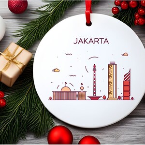 Könnte beinhalten: Weißer Keramik-Anhänger mit roter Schnur und dem Wort "JAKARTA" in roten Buchstaben. Der Anhänger zeigt eine Linienzeichnung der Skyline von Jakarta in Rot und Gelb.