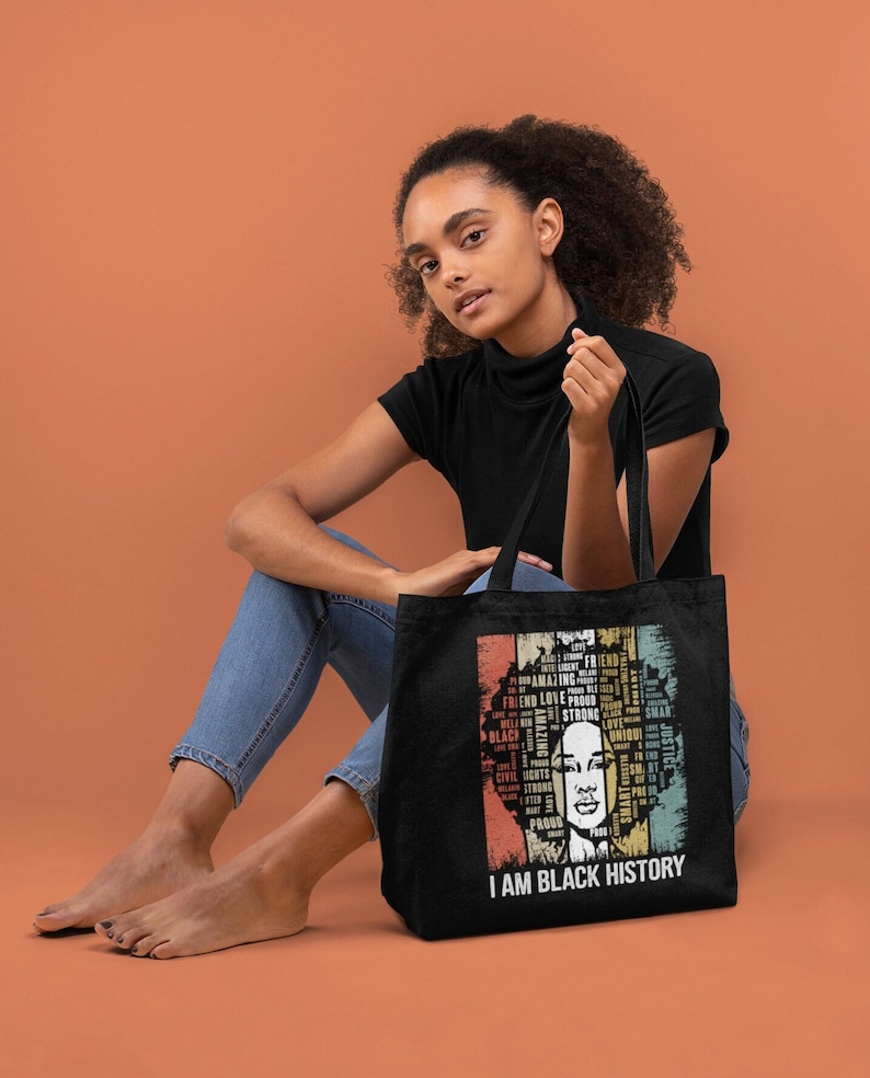 Puede incluir: Una bolsa de tela negra con un gr&aacute;fico de estilo vintage de la cara de una mujer formada por palabras como "amigo", "amor", "fuerte" y "negro". El texto "Soy historia negra" est&aacute; impreso debajo del gr&aacute;fico.