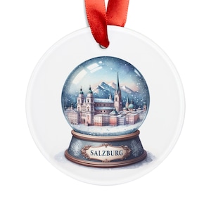 May include: A white ceramic ornament shaped like a snow globe with a blue base and a gold trim. The snow globe depicts a snowy cityscape with a church and other buildings. The base of the snow globe has the word "SALZBURG" written on it.