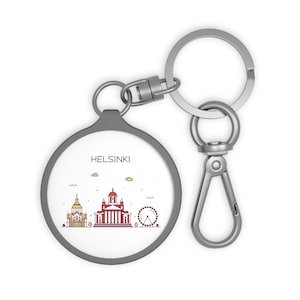 May include: A silver keychain with a round, gray charm featuring a line drawing of the Helsinki skyline. The charm has the word "HELSINKI" printed in black at the top.