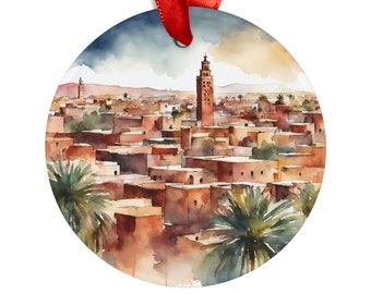 Op de afbeelding: Een aquarel schilderij van een stadsgezicht met een hoge minaret in het midden. De gebouwen zijn geschilderd in tinten bruin en oranje, en de lucht is een zachte blauwe kleur met witte wolken. Het schilderij is ingelijst in een rond, metalen ornament.