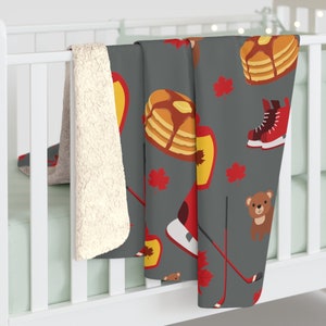 May include: A gray fleece blanket with a Canadian themed pattern. The pattern includes maple leaves, hockey sticks, a bear, and stacks of pancakes.