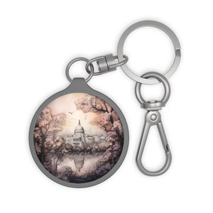 May include: A silver keychain with a round, gray metal charm featuring a watercolor illustration of a building with a dome, surrounded by trees and a reflection in water.