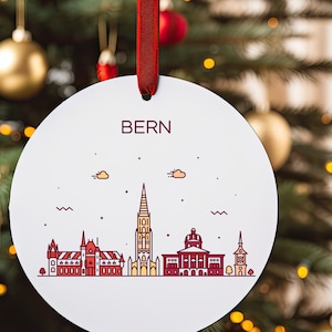 Puede incluir: Adorno redondo blanco con una cinta roja, que presenta un dibujo lineal del horizonte de Berna con la palabra "BERN" en letras rojas.