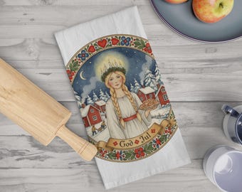 May include: White kitchen towel with a watercolor illustration of a woman with a candle crown, holding pastries, and the text "God Jul". The design includes red houses, snow, and a banner with floral accents.