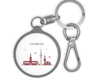 May include: A silver key chain with a round, grey charm featuring a line drawing of the Hamburg skyline. The charm has the word "HAMBURG" printed in black at the top.