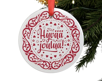 Könnte beinhalten: Ein weißer, runder Weihnachtsschmuck mit rotem Zierrand und dem finnischen Text "Hyvää joulua!" (Frohe Weihnachten). Der Schmuck hängt an einem roten Band und vor einem Weihnachtsbaum.