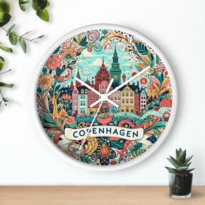 May include: White round clock with a colorful illustration of Copenhagen, Denmark. The clock face features a stylized cityscape with buildings, water, and floral details. The text "Copenhagen" is written in a banner at the bottom.