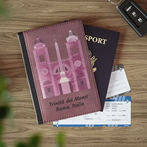 May include: A pink passport holder with a graphic of the Trinità dei Monti church in Rome, Italy. The text "Trinità dei Monti Roma, Italia" is printed on the front of the holder.