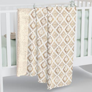May include: A soft, plush throw blanket with a white background and a geometric pattern of brown squares. The blanket is folded and draped over a white crib.