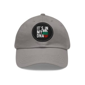 Op de afbeelding: Grijze baseballpet met een gebogen klep. De pet heeft een ronde patch met de tekst "IT'S IN MY DNA" en een afbeelding van de Bulgaarse vlag in groen, wit en rood.