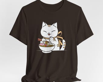 May include: A dark brown t-shirt featuring a white cat illustration. The cat is depicted with a yellow bow, holding chopsticks and eating ramen from a white bowl. The ramen includes noodles, meat, and vegetables.