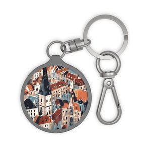 Op de afbeelding: Een zilveren sleutelhanger met een ronde, zilveren hanger met een kleurrijke illustratie van een stadsbeeld. De hanger heeft een zilveren rand en het stadsbeeld is geschilderd in tinten rood, oranje, blauw en wit.